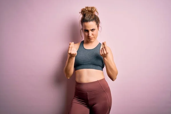 Young beautiful blonde sportswoman doing sport wearing sportswear over pink background doing money gesture with hands, asking for salary payment, millionaire business