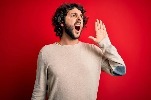 髭を生やした若いハンサムな男が赤い背景に立ってカジュアルなセーターを着て大声で叫び 口の中で手で横に大声で叫ぶ コミュニケーションの概念 — ストック写真