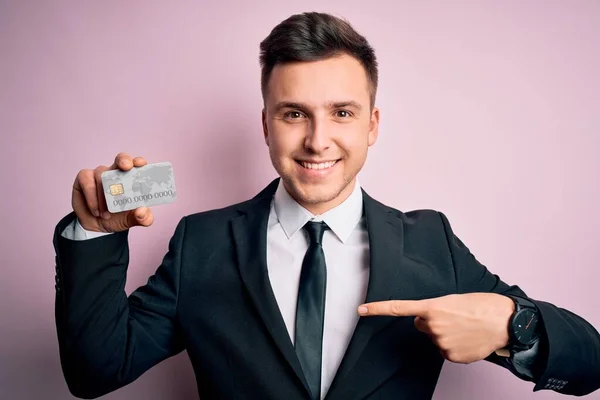 Young handsome caucasian business man holding finance credit card over pink background with surprise face pointing finger to himself