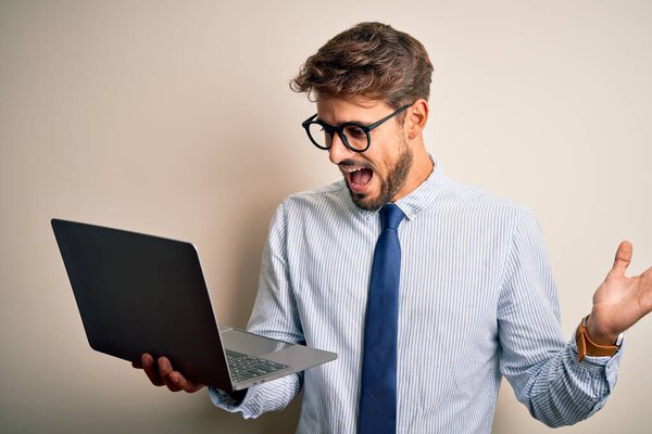 Молодой бизнесмен в очках, работающий с ноутбуком на белом фоне, очень счастлив и взволнован, выражение лица победителя, празднующего победу, кричит с большой улыбкой и поднимает руки