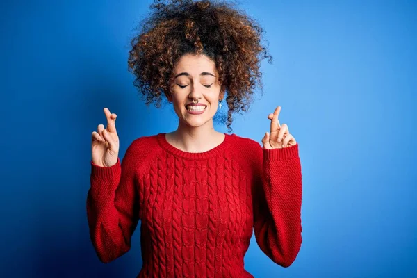 Young beautiful woman with curly hair and piercing wearing casual red sweater gesturing finger crossed smiling with hope and eyes closed. Luck and superstitious concept.