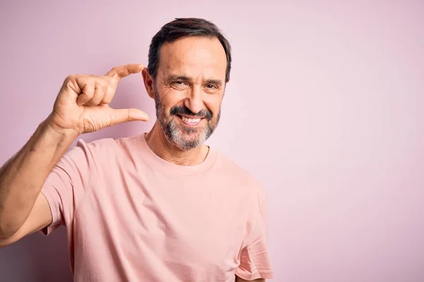 孤立したピンクの背景の上に立っているカジュアルなTシャツを身に着けている中年のホーリーマンは 指を見てカメラと小さなサイズの看板を行う手で笑顔と自信を持ってジェスチャー 概念の測定 — ストック写真