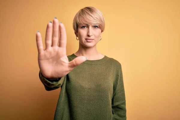 Young blonde woman with modern short hair wearing casual sweater over yellow background doing stop sing with palm of the hand. Warning expression with negative and serious gesture on the face.