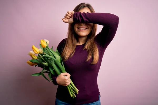 Young blonde woman holding romantic bouquet of yellow tulips flowers over pink background Smiling cheerful playing peek a boo with hands showing face. Surprised and exited