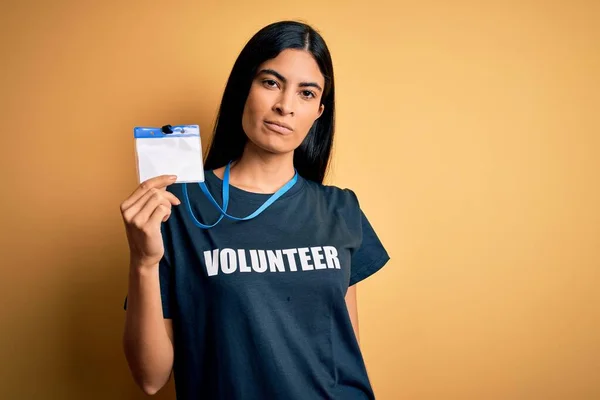 Young beautiful hispanic woman wearing volunteer t-shirt and showing identification with a confident expression on smart face thinking serious