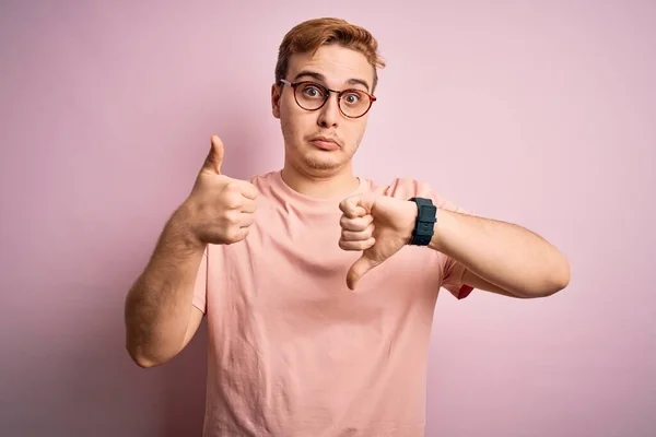 Young handsome redhead man wearing casual t-shirt standing over isolated pink background Doing thumbs up and down, disagreement and agreement expression. Crazy conflict