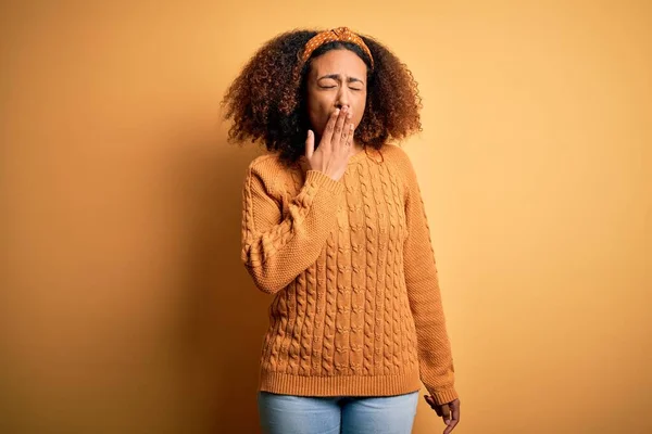 Young african american woman with afro hair wearing casual sweater over yellow background bored yawning tired covering mouth with hand. Restless and sleepiness.