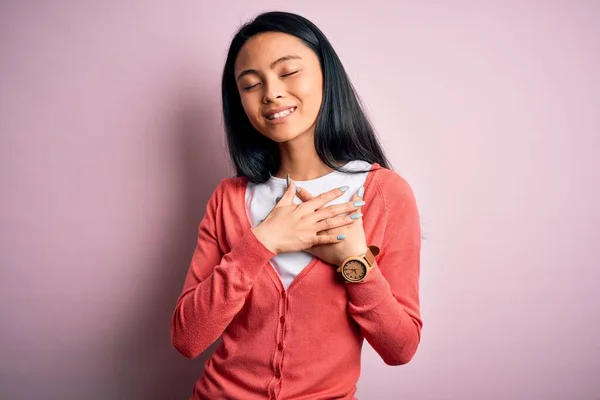 孤立したピンクの背景の上にカジュアルなセーターを着て若い美しい中国の女性は 目を閉じて胸の上に手で笑顔と顔に感謝のジェスチャー 健康コンセプト — ストック写真