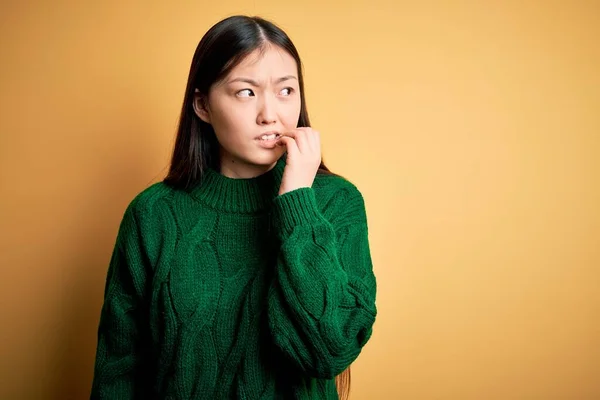 年轻美丽的亚洲女人穿着绿色冬季毛衣 披着黄色孤立的背景 看起来紧张而紧张 双手放在咬指甲的嘴上 焦虑问题 — 图库照片