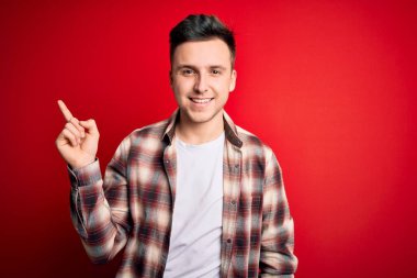Kırmızı izole arka plan üzerine gündelik modern gömlek giyen yakışıklı beyaz bir adam. Yüzünde büyük bir gülümseme, kameraya el parmağı ile bakıyor..