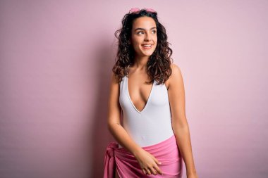 Tatilde kıvırcık saçlı güzel bir kadın pembe arka planda beyaz mayo giyip yüzünde gülümseme, doğal bir ifade ile bir o yana bir bu yana bakıyor. Kendinden emin gülen.