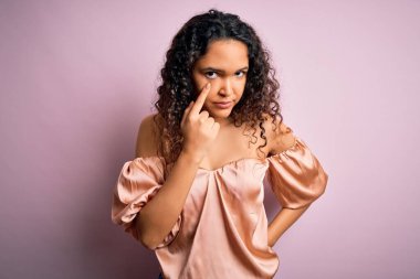 Kıvırcık saçlı, pembe tişörtlü genç ve güzel bir kadın pembe arka planda dikiliyor.