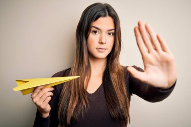Genç, güzel bir kız kağıttan bir uçak tutuyor, beyaz arka planda duruyor, açık elle dur işareti yapıyor, ciddi ve kendinden emin bir ifadeyle, savunma jesti yapıyor.