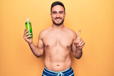 Tatilde sakallı yakışıklı bir adam mayo giyiyor elinde bir şişe aloe vera kremi bir fikir veya soruyla gülümsüyor.