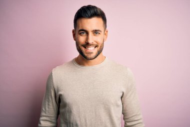 Yüzünde mutlu ve havalı bir gülümsemeyle pembe bir arka plan üzerinde duran sıradan bir süveter giyen genç yakışıklı bir adam. Şanslı kişi..