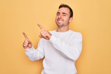 Mavi gözlü, sıradan bir süveter giyen, sarı arka planda duran, gülümseyen ve kameraya bakan, iki eliyle ve parmaklarıyla yüzünü gösteren genç yakışıklı bir adam..