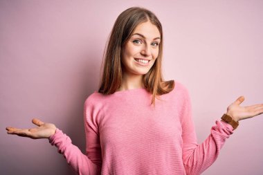 Güzel, kızıl saçlı bir kadın pembe arka plan üzerine günlük süveter giyip gülümseyerek avuçlarını açıyor, sunuyor ve reklam karşılaştırması yapıyor ve dengeyi sağlıyor.
