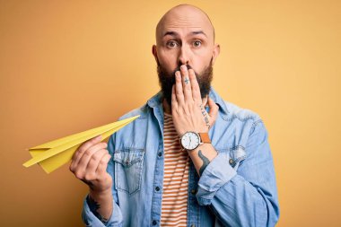 Kel ve sakallı yakışıklı bir adam, sarı arka planda kâğıt uçak tutuyor. Ağzı kapalı, utancından ağzı açık, korkudan ağzı açık kalmış, sessizlikten korkan, gizli bir konsept.