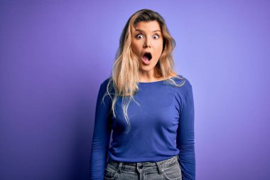 Genç, güzel, sarışın bir kadın, izole edilmiş mor arka plan üzerine gündelik tişört giyip şaşkınlık ve şaşkınlık dolu bir ifade, korku ve heyecanlı bir yüz ifadesi ile şoka girmiş..