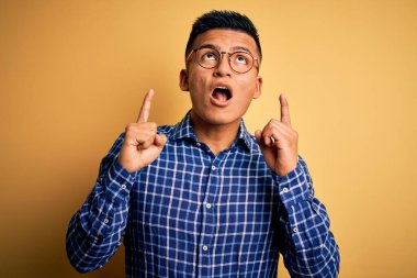 Sarı arka planda gündelik gömlek ve gözlük takan genç, yakışıklı latin adam şaşırmış ve şaşırmış bir şekilde yukarı bakıp parmaklarını göstermiş ve kollarını kaldırmıştı..
