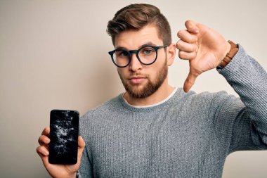 Sakallı, mavi gözlü, gözlüklü, kızgın yüzlü, kırık akıllı telefon takan, olumsuz işaretli, baş parmakları aşağıda, reddedilme konsepti olan genç sarışın bir adam.