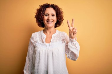 Orta yaşlı güzel kıvırcık saçlı kadın sarı arka planda rahat bir yaz elbisesi giyiyor ve kameraya gülümsüyor zafer işareti yapıyor. İki numara..