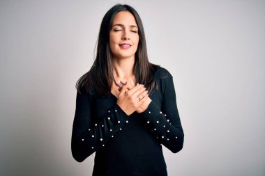 Mavi gözlü genç esmer kadın beyaz arka planda siyah bir süveter giyiyor elleri göğsünde, gözleri kapalı ve yüzünde minnet dolu bir jest ile gülümsüyor. Sağlık kavramı.