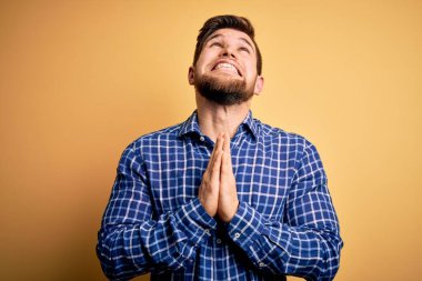 Sarışın, sakallı ve mavi gözlü genç bir işadamı sarı arka planda tişört giyiyor. Elleriyle yalvarıyor ve dua ediyor. Yüzünde umut ifadesi var. Yalvarıyorum..