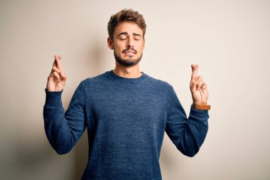 Sakallı, sıradan bir süveter giyen, beyaz arka plan jestleri yapan, umut dolu ve gözleri kapalı gülümseyen genç yakışıklı bir adam. Şans ve batıl inanç..