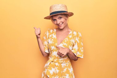 Sarı arka planda sarı şapkalı, tatilde olan güzel sarışın bir kadın. Arka tarafı işaret ediyor. El ve baş parmakları havada, kendine güvenen.