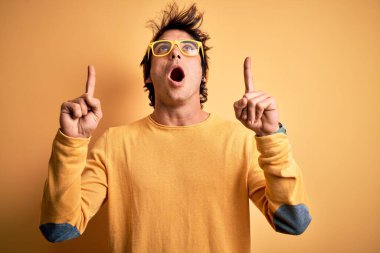 Sarı arka plan üzerinde gündelik tişört ve gözlük takan genç yakışıklı adam şaşırmış ve şaşırmış ve parmaklarıyla işaret etmiş ve kollarını kaldırmış..