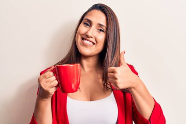 Genç, güzel esmer kadın bir kupa kahve içiyor mutlu ve pozitif gülümsüyor, başparmağıyla mükemmel ve onay işareti yapıyor. 
