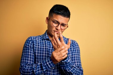 Sarı arka planda gündelik gömlek ve gözlük takan genç, yakışıklı Latin adam. Ellerinde ve parmaklarında ağrı, iltihaplanma.