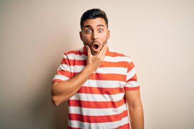 İzole edilmiş beyaz arka plan üzerinde duran sıradan çizgili tişört giyen genç yakışıklı adam, elleri çenesinde olan inançsızlıktan, şaşkınlıktan ve şaşkınlıktan büyülenmiş görünüyor.