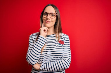 Genç, mavi gözlü, gözlüklü, kırmızı arka planda duran ve kameraya kendinden emin, kollarını çaprazlayarak gülümseyen ve çenesini kaldıran güzel bir sarışın kadın. Olumlu düşünüyorum..