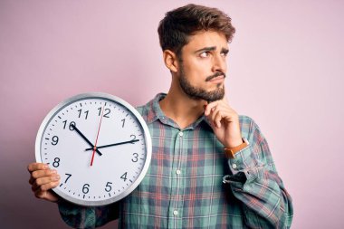 Sakallı genç adam geri sayım yapıyor. Tecrit edilmiş pembe arka plan üzerinde büyük bir saat kullanıyor. Ciddi bir yüz.