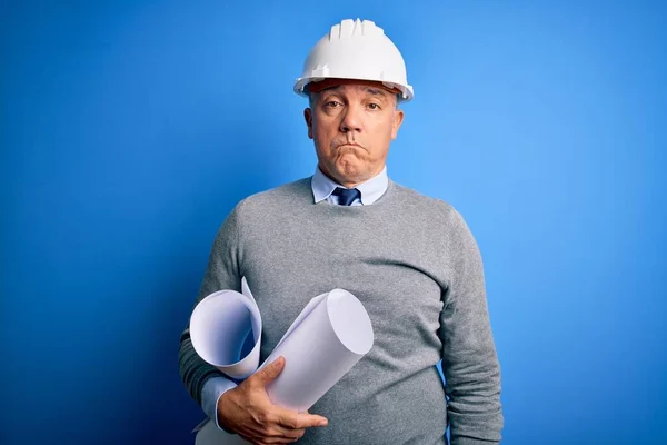 Middle age handsome grey-haired architect man wearing safety helmet holding blueprints depressed and worry for distress, crying angry and afraid. Sad expression.