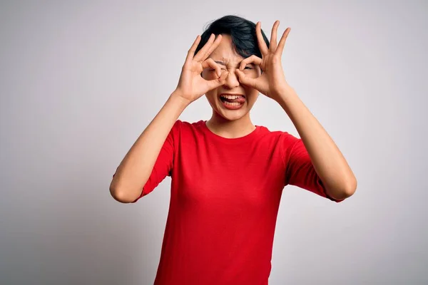 年轻美丽的亚洲女孩穿着休闲的红色T恤 站在孤立的白色背景上 做着像双筒望远镜一样伸出舌头的动作 眼睛透过手指看着 疯狂的表情 — 图库照片