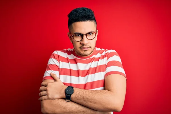 Young handsome man wearing casual striped t-shirt and glasses over isolated red background shaking and freezing for winter cold with sad and shock expression on face