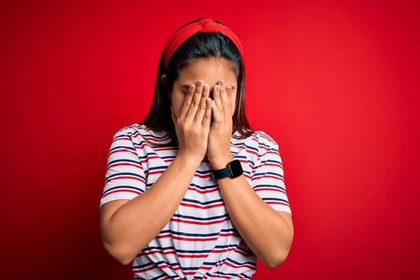 年轻美丽的亚洲女孩穿着宽松的条纹T恤 披着孤立的红色背景 脸上挂着忧郁的表情 一边哭一边用手捂住脸 抑郁症的概念 — 图库照片