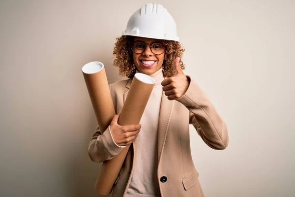 非洲裔美国妇女 卷曲的建筑师 戴着安全帽 戴着眼镜 手里拿着设计图 带着大大的笑容 高兴地做着好的手势 用手指竖起大拇指 很好的标志 — 图库照片