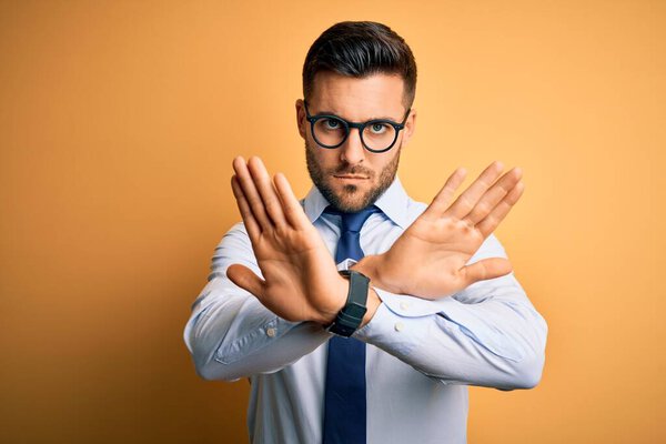 Young handsome businessman wearing tie and glasses standing over yellow background Rejection expression crossing arms doing negative sign, angry face