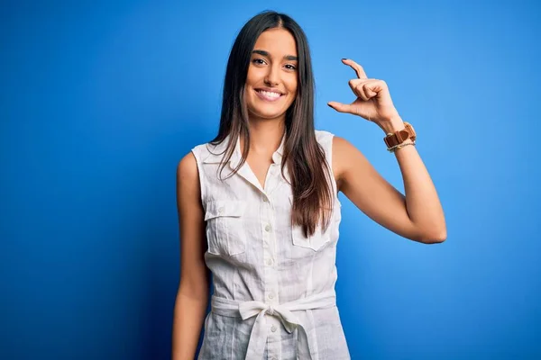 Young beautiful brunette woman wearing casual dress over isolated blue background smiling and confident gesturing with hand doing small size sign with fingers looking and the camera. Measure concept.