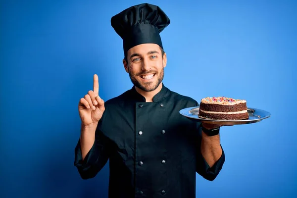 ひげを生やした若いハンサムな炊飯器男の制服と帽子を保持トレイでケーキ驚きでアイデアや質問を指差す指で幸せな顔 ナンバーワン — ストック写真