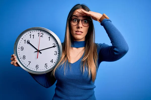 Young blonde woman with blue eyes holding big minute clock over isolated background stressed with hand on head, shocked with shame and surprise face, angry and frustrated. Fear and upset for mistake.