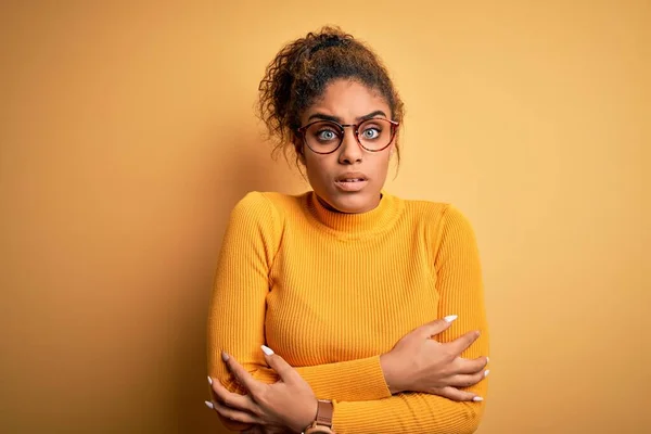 Young beautiful african american girl wearing sweater and glasses over yellow background shaking and freezing for winter cold with sad and shock expression on face