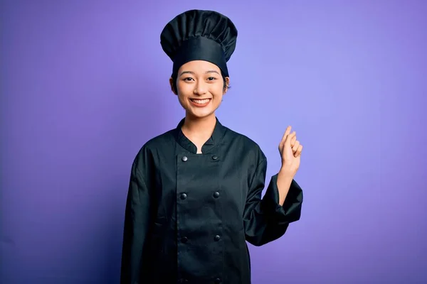 年轻美丽的中国厨师女士身穿炊具制服 头戴紫色背景的帽子 脸上挂着大大的笑容 手指头指向旁边看相机 — 图库照片