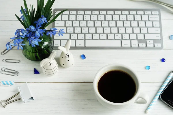 Easter concept. Flat lay office table feminine desk. workspace with laptop, phone, bouquet blue flowers, figures of hares on a white wooden background.