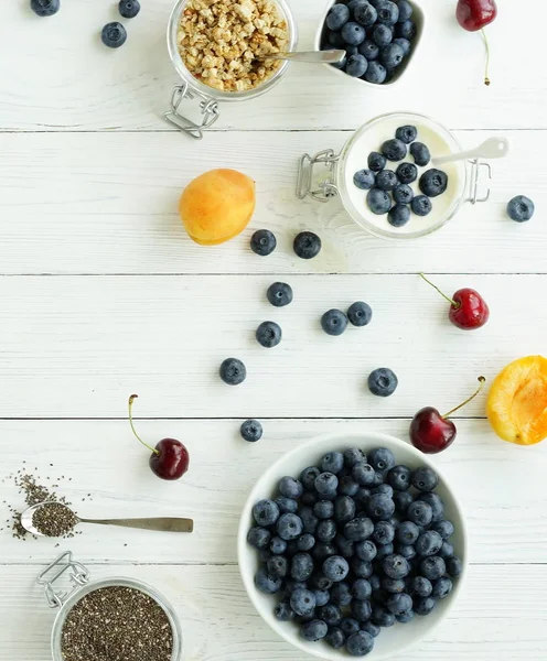 En god och hälsosam frukost med färska blåbär, granulat, chiafrön, yoghurt med blåbär i en glasburk på en vit trä bakgrund. Ovanifrån — Stockfoto