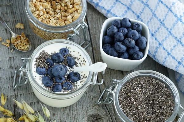 En god och hälsosam frukost med färska blåbär, granulat, chiafrön, yoghurt med blåbär i en liten glasburk på en vintage trä bakgrund. Ovanifrån — Stockfoto